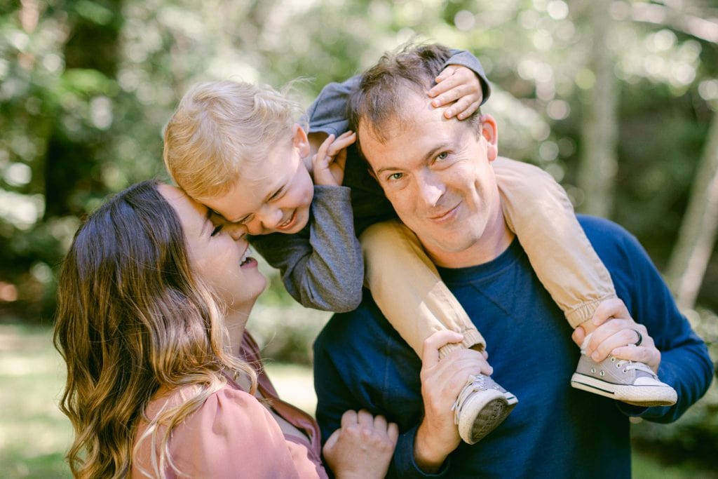 Father holding son on shoulders while mother comes in to kiss him.
