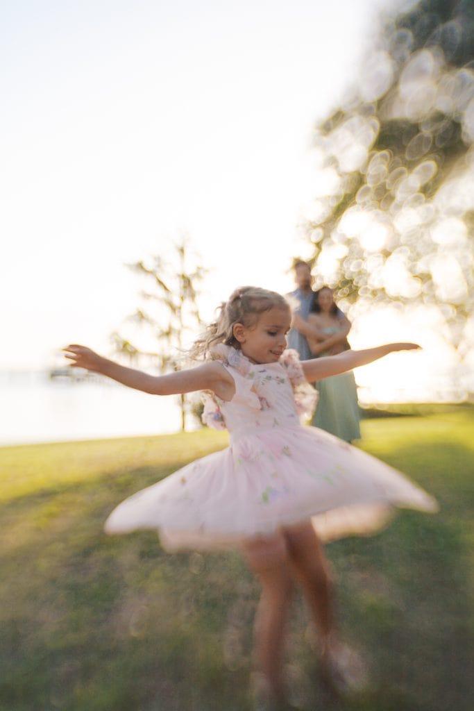 Young girl in a pink tutu spinning in a grassy field with sunlight filtering through trees.